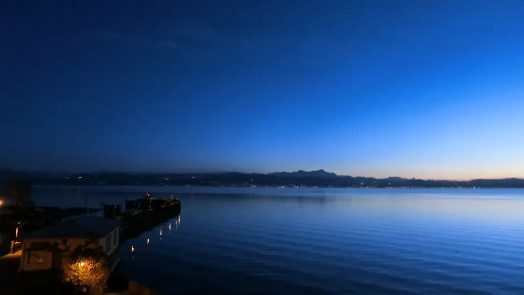 Blaue Stunde an einem See, vorn ein beleuchteter Steg, am Horizont Berge