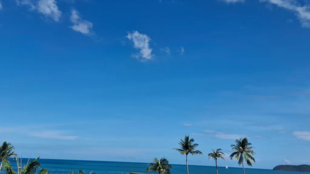 Blauer Himmel mit Wolken über dem Meer, am Ufer einzelne Palmen